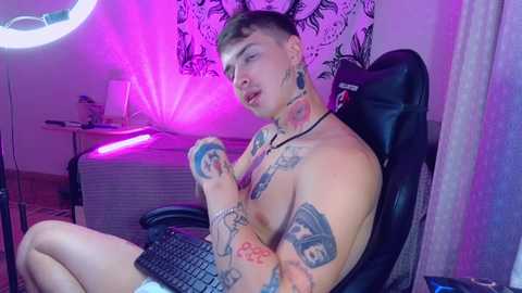 Media: Video of a topless, tattooed, light-skinned male with short, dark hair, sitting at a gaming chair, typing on a keyboard, in a dimly lit room with a colorful, patterned tapestry and purple LED lighting.