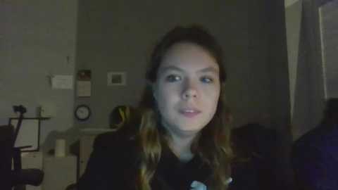 Media: A video of a young woman with light skin and long, wavy brown hair, wearing a black top, standing in a dimly lit room with a grey wall, a white clock, and a black tripod in the background.