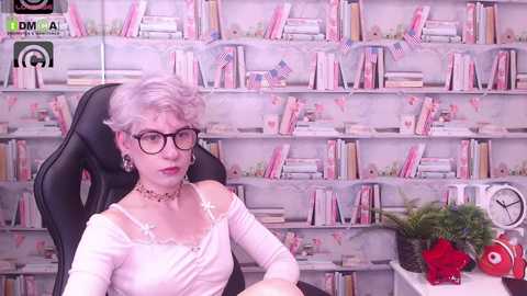 Media: Video of a white woman with short, platinum blonde hair, wearing glasses, a white off-shoulder top, and a choker, seated in a black chair in a room with pink bookshelves filled with books and a clock.