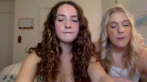 Media: Video of two young women with curly hair, one with brown and the other with blonde, both with closed eyes, smiling. They are in a bedroom with a white door and a framed picture.