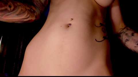 Media: Video of a fair-skinned woman with a pierced navel and tattooed arms, viewed from the side, revealing her bare torso against a dark background.