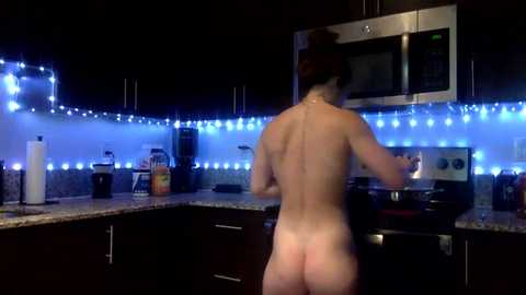 Media: A video of a nude woman with auburn hair, standing in a dimly lit kitchen with blue LED lights, preparing a meal on the stove.