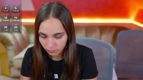 Media: A video of a young woman with long dark hair, wearing a black shirt, seated indoors with a blurred background featuring a red wall and a blue chair.