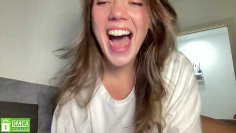 Media: Video of a young woman with long, wavy brown hair, wearing a white shirt, laughing wildly. Background features a grey couch and a white wall with a door slightly ajar.