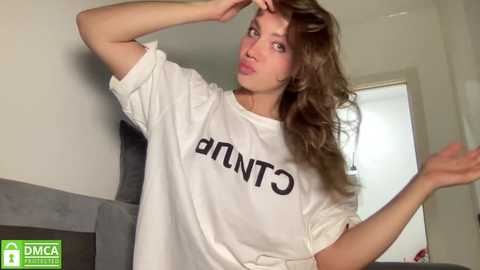 Media: Video of a young Caucasian woman with wavy brown hair, wearing a white oversized T-shirt with \"CUNT\" in bold black letters. She has a playful expression, posing in a modern, minimally decorated room with grey couches and white walls.