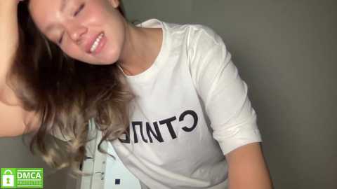 Media: A video of a young Caucasian woman with wavy, shoulder-length brown hair, wearing a white T-shirt with the word \"GENT\" in bold black letters. She is smiling, leaning forward, with her left hand in her hair.