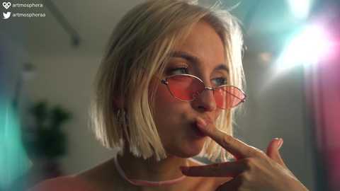 Media: Video of a blonde woman with short hair, wearing pink glasses, making a peace sign with her fingers. She has a light skin tone and is in a dimly lit room with blurred background.