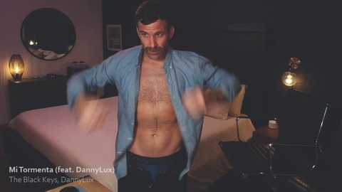 Media: Video of a shirtless, muscular man with dark hair and a beard, wearing an unbuttoned blue shirt, in a dimly lit room with a bed, nightstand, and lamp.