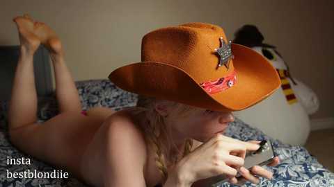 Media: Video of a nude woman with blonde hair, wearing an orange cowboy hat, lying on a bed with blue and white patterned sheets, reading a book.