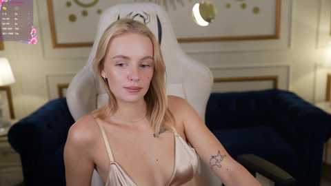 Media: Video of a young, slender, blonde woman with fair skin, wearing a low-cut beige lace bralette, sitting on a plush gaming chair in a dimly lit, cozy room.