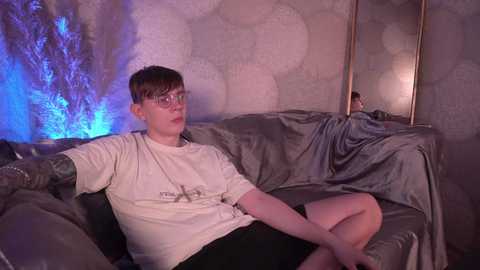 Media: A young person with short brown hair, glasses, and a light t-shirt sits on a gray couch, draped in a silver sheet, with a blue light illuminating the background.