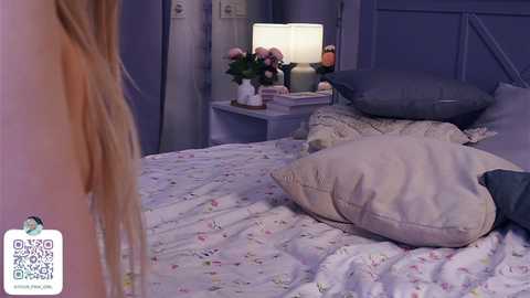 Media: Video of a dimly-lit bedroom with a blonde woman's back to the camera, a floral bedspread, and a nightstand with a lamp, flowers, and books.