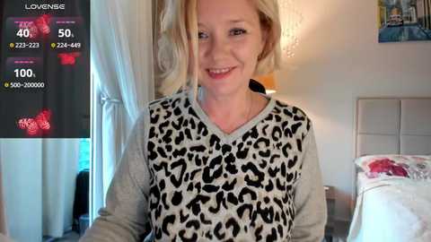 Media: Video of a smiling blonde woman in a leopard print sweater, standing in a cozy bedroom with a bed and lamp, with a Lovense app interface on the left.