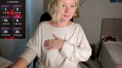 Media: Video of a blonde woman with shoulder-length hair wearing a loose white top, sitting on a black chair in a dimly lit bedroom.