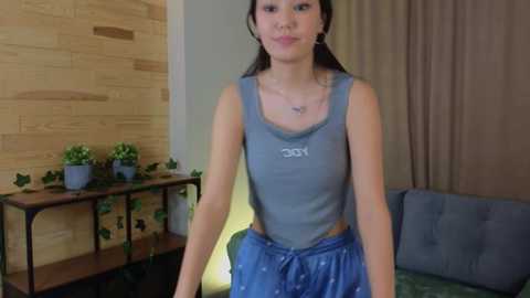 Media: Video of a young Asian woman with long dark hair in a gray tank top and blue pajama pants, standing in a modern living room with wooden walls, green plants, and a gray couch.