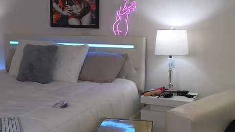 Media: A video of a modern, minimalist bedroom with a white bed, a neon pink figure silhouette on the wall, a white lamp, and a white side table.