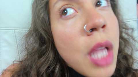 Media: Video of a young woman with curly brown hair, light skin, and a silver nose ring, wearing a black top, with a surprised expression.