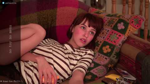 Media: Video of a young woman with short brown hair, wearing a striped dress, reclining on a colorful patchwork sofa.