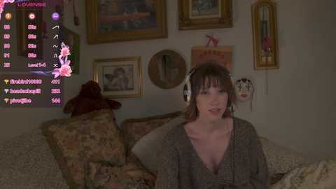 Media: Video of a young woman with fair skin, medium-length brown hair, and large headphones, sitting on a bed with patterned bedding in a cozy, dimly-lit room adorned with various framed paintings and decorations.