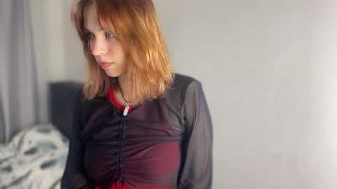 Media: Video of a young woman with shoulder-length, straight, reddish-brown hair, wearing a black, sheer blouse with a red lining, looking contemplative against a gray wall.