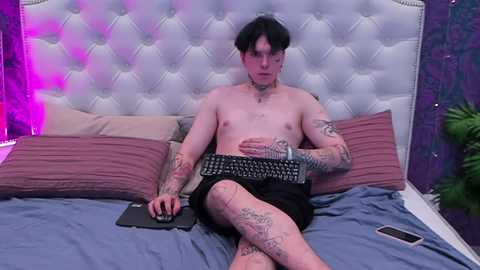 Media: Video of a shirtless, tattooed, pale-skinned man with black hair, wearing black shorts, sitting on a bed with a tufted white headboard, typing on a keyboard, surrounded by pillows and a smartphone.