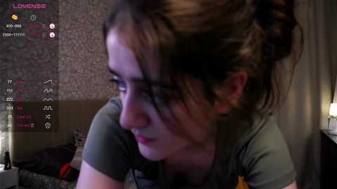 Media: A dimly lit video of a young woman with fair skin and brown hair tied in a bun, wearing glasses and a gray shirt, leaning forward. Background includes a floral wallpaper and a nightstand with a lamp.