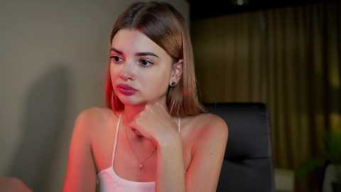 Media: Video of a young, light-skinned woman with straight, shoulder-length brown hair, wearing a pink spaghetti-strap top, sitting indoors, with a shadowy background and red lighting.