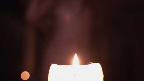 Media: Video of a lit candle in a dark, blurry room, casting a warm, glowing light and creating soft shadows. The candle flame is bright and steady, illuminating the edges of the wick and the surrounding wax.