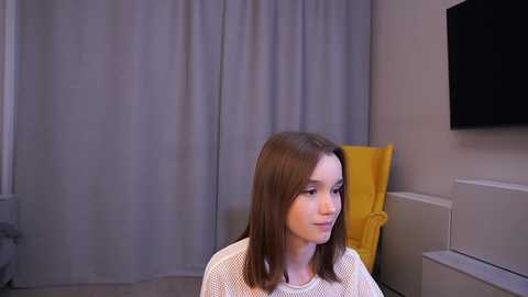 Media: Video of a young girl with straight, shoulder-length brown hair, wearing a white knit sweater, seated indoors. Background features gray curtains, a yellow cushion, and a flat-screen TV mounted on a beige wall.