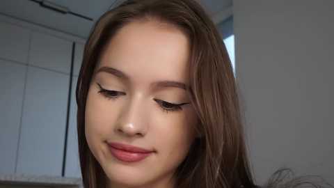 Media: Video of a young, light-skinned woman with straight, shoulder-length brown hair, wearing minimal makeup, and a soft, closed-mouth smile. The background shows a modern, minimalist room with white walls and a closet.