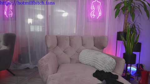 Media: Video of a cozy, dimly-lit room with a plush, pink chaise lounge, white throw blanket, and a tall, green plant. Neon signs with nude silhouettes hang on the wall, and purple and white lighting creates a sensual ambiance.