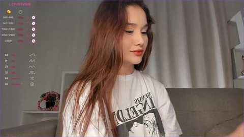 Media: A video of a young woman with long, straight brown hair, wearing a white t-shirt with a graphic design, sitting in a modern living room with gray furniture.