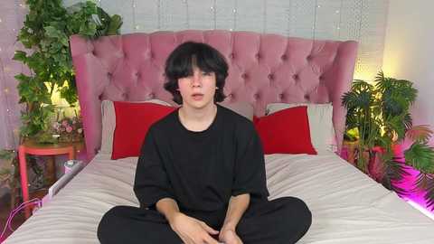 Media: Video of a young woman with short black hair, wearing a black sweatshirt, sitting cross-legged on a bed with a pink tufted headboard, surrounded by green plants and red pillows, in a cozy, well-lit room.