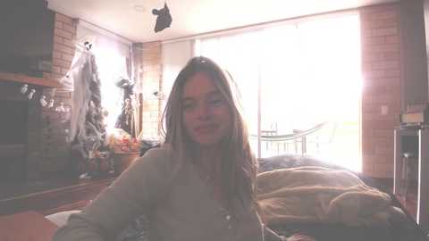 Media: Video of a young woman with long, straight brown hair, wearing a light-colored shirt, sitting on a bed in a dimly lit room with brick walls, Halloween decorations, and a large window.