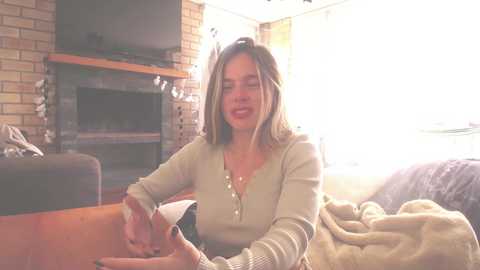 Media: Video of a smiling woman with light brown hair in a beige sweater, sitting in a cozy living room with a fireplace, brick wall, and natural light streaming through large windows.