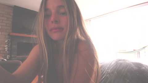 Media: Video of a topless young woman with long, straight blonde hair, seated indoors near a window. Brick fireplace and a green hammock visible in the background.