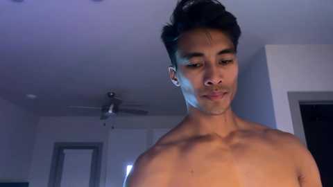 Media: Video of a shirtless, fit young man with short black hair, tan skin, and a slight smile, standing indoors with white walls and a ceiling fan visible in the background.