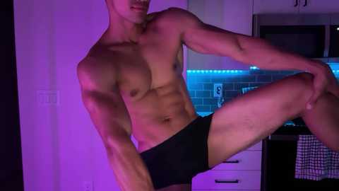 Media: Video of a muscular, shirtless man with light skin wearing black briefs, standing in a modern kitchen with purple lighting and a tiled backsplash.