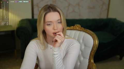 Media: A video of a blonde woman with fair skin and blue eyes, wearing a white blouse, contemplatively biting her finger in a modern living room with a green couch and gold-framed mirror.