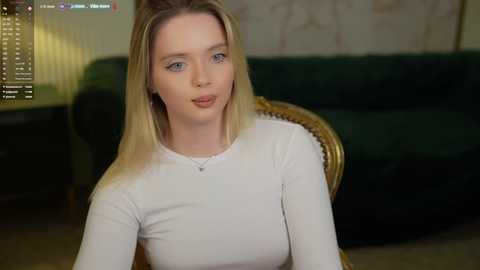 Media: Video of a young, fair-skinned woman with straight blonde hair, wearing a white long-sleeved top, sitting indoors. Background features a green sofa, a dimly lit room, and a TV screen displaying a video feed.