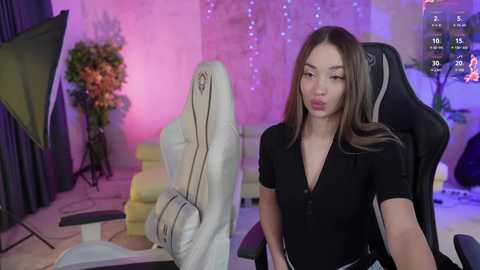 Media: Video of a young Asian woman with long brown hair, wearing a black V-neck top, sitting in a black gaming chair, in a colorful, well-lit room with a desk, plants, and fairy lights.