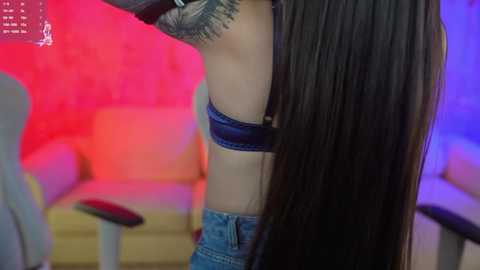 Media: Video of a woman with long, straight, dark hair, wearing a blue bra and denim shorts, standing in a dimly lit room with pink and purple lights.