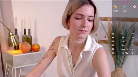 Media: Video of a light-skinned woman with a bob haircut, wearing a sleeveless white top, in a minimalist room with modern decor, including a table with wine bottles, a lamp, and a vase.