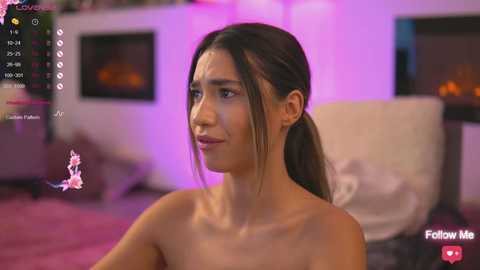 Media: Video of a topless, young, light-skinned Latina woman with long dark hair in a ponytail, sitting indoors with a serene expression. Background features a fireplace and soft lighting.