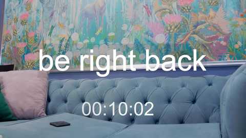 Media: Video of a pastel-colored, tufted sofa with a smartphone on it, against a vibrant, floral-patterned mural wall. Text reads \"be right back 00:10:02.\