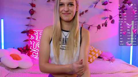 Media: Video of a young woman with long blonde hair, wearing a white tank top, standing in a bedroom with pink and purple lights, plush toys, and a calendar on the wall.