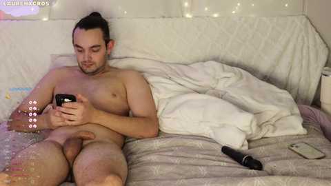 Media: A video of a nude man with a circumcised penis, lying on a bed, holding a phone, surrounded by a messy room with white sheets and scattered items.