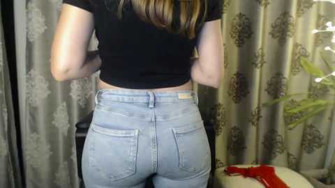 Media: Video of a woman from behind, wearing a black crop top and high-waisted light blue jeans, showing off her curvy, muscular buttocks. She stands in a room with floral-patterned curtains and a potted plant.