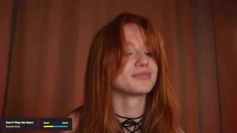 Media: Video of a smiling young woman with long, red hair, wearing a black choker, set against a warm-toned, blurred background.