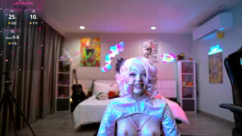 Media: Video of a woman in a shiny, metallic, revealing outfit, standing in a colorful, modern bedroom with a bed, colorful wall art, and pink heart balloons.